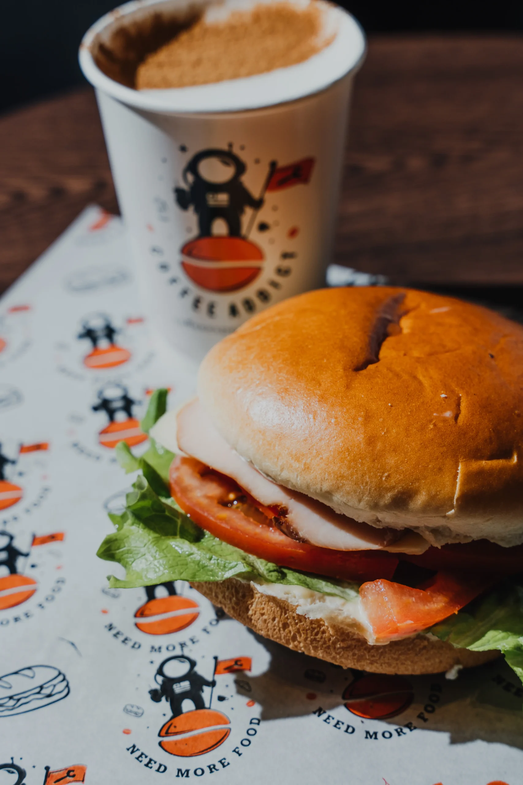Close-up of a hamburger with lettuce and tomato in a soft bun, foreground; a space-theme coffee cup sits blurred in the background.