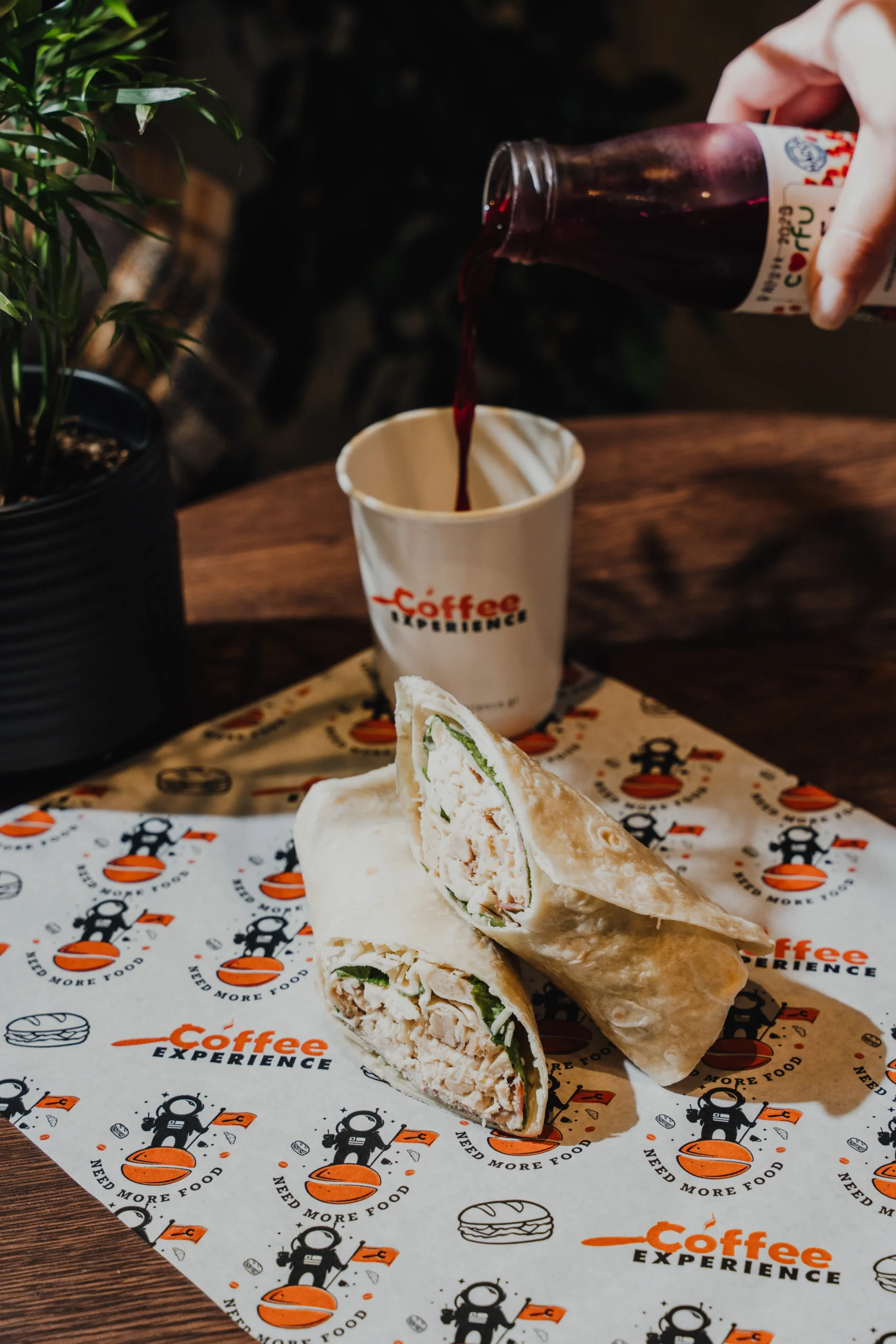 Hand pours a dark red drink from a bottle into a white cup labeled Coffee Experience, with wrapped sandwiches on the table.
