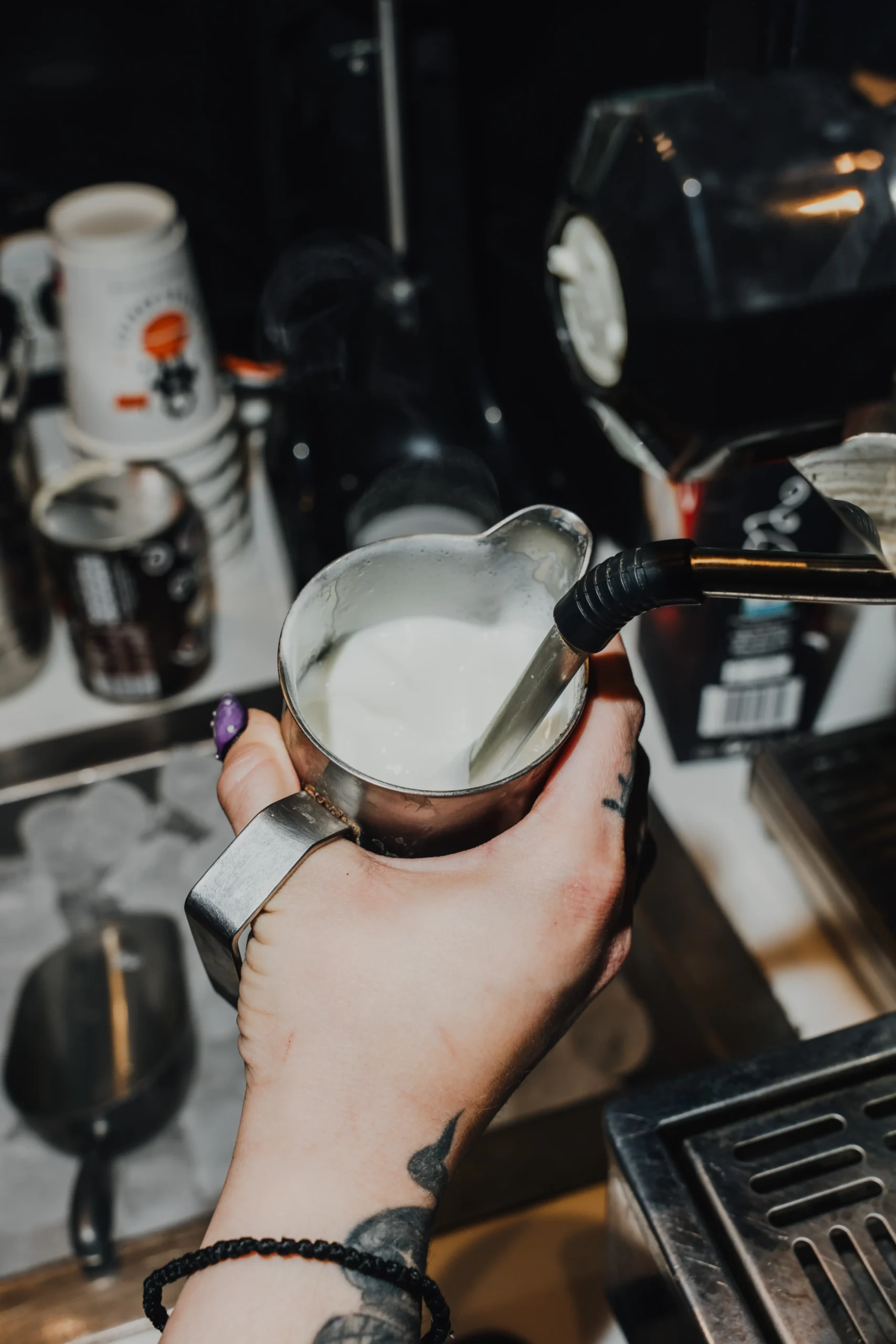 Person's hands holding a metal pitcher of milk for frothing in front of a espresso machine in a cafe.